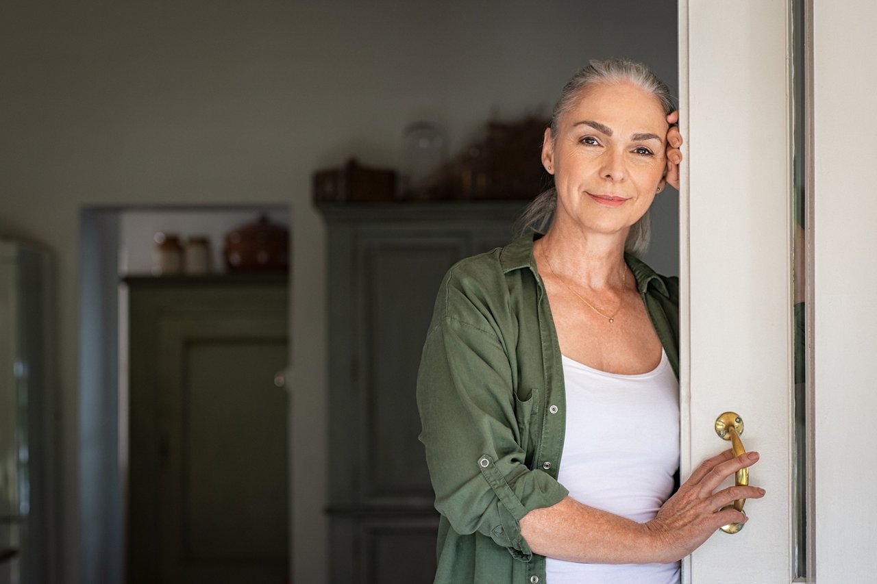 Portrait of senior woman standing while leaning against door at home and looking at camera. Smiling mature woman standing at doorway and looking at camera. Portrait of old lady relaxing and thinking with copy space.